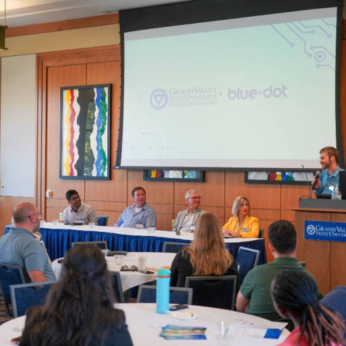 A speaker stands at a podium addressing an audience during a panel discussion at Grand Valley State University, with five panelists seated at a table on stage. The projector screen behind them displays the logos of the GVSU College of Computing and Blue D
